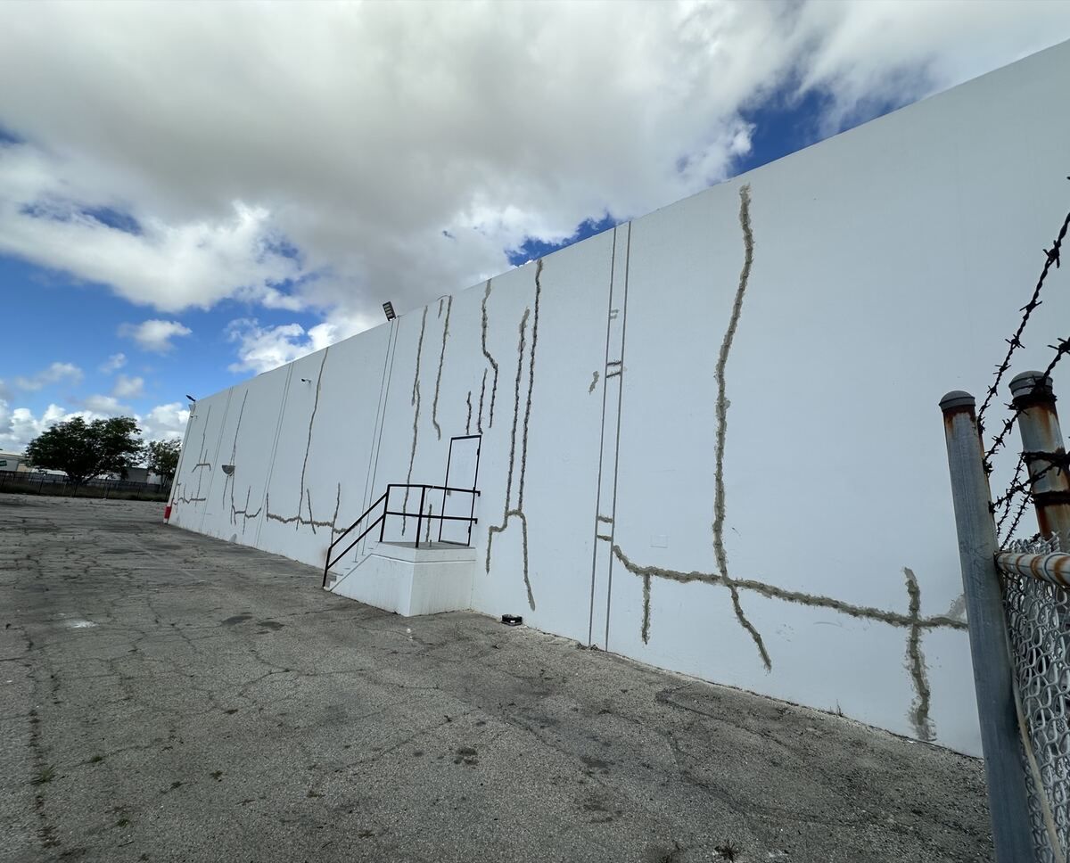Long cracks sealed with grey paste running down a white tilt-up wall beside an exterior steel service stair