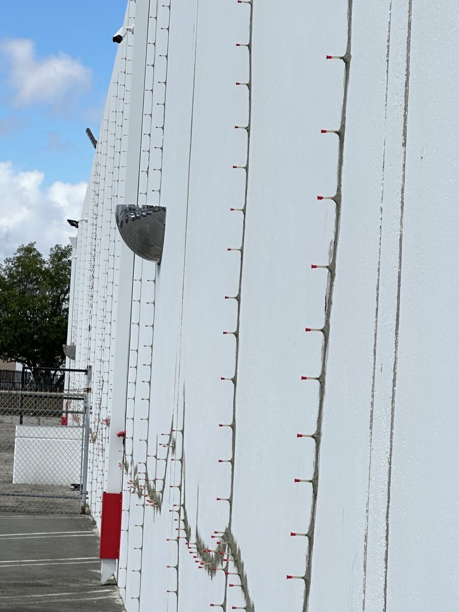 Tilt-up concrete wall seen at an angle along its face, with vertical cracks surface-sealed and lined with red-capped epoxy injection ports running from the top of the wall to the ground