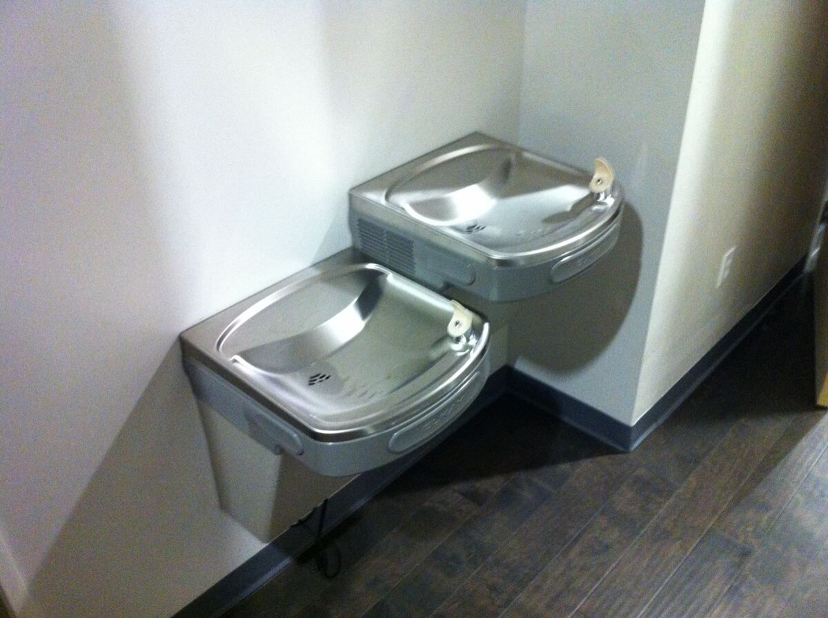 Two stainless steel drinking fountains mounted at two different heights in a corner above a dark wood-plank floor