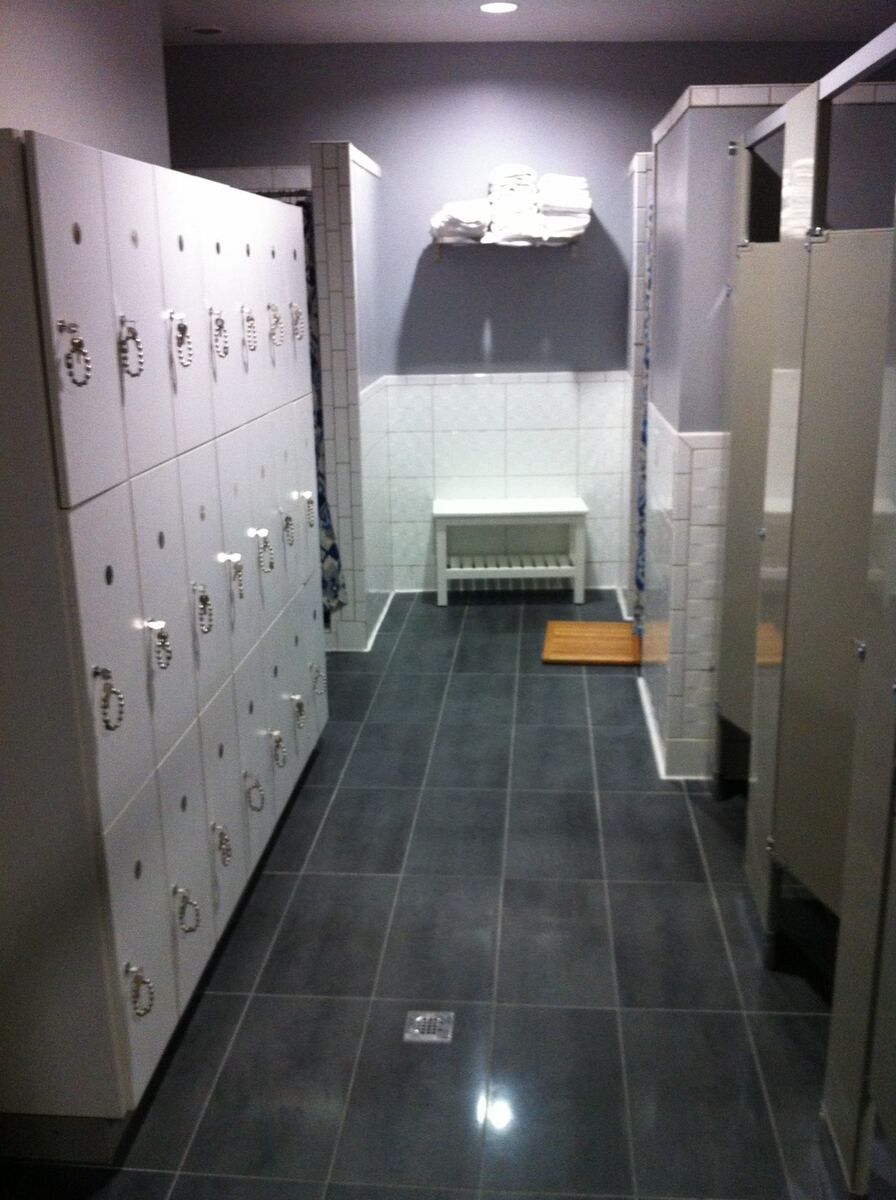Finished locker room with a wall of lockers on the left, tiled shower and toilet partitions on the right, a bench in a white-tiled alcove at the back and a dark grey tile floor