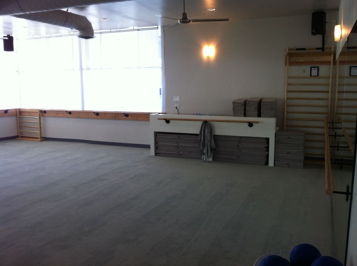 Finished studio with a long wall-mounted barre under a shaded window, exposed round ductwork and ceiling fans overhead, and a built-in bench stacked with mats